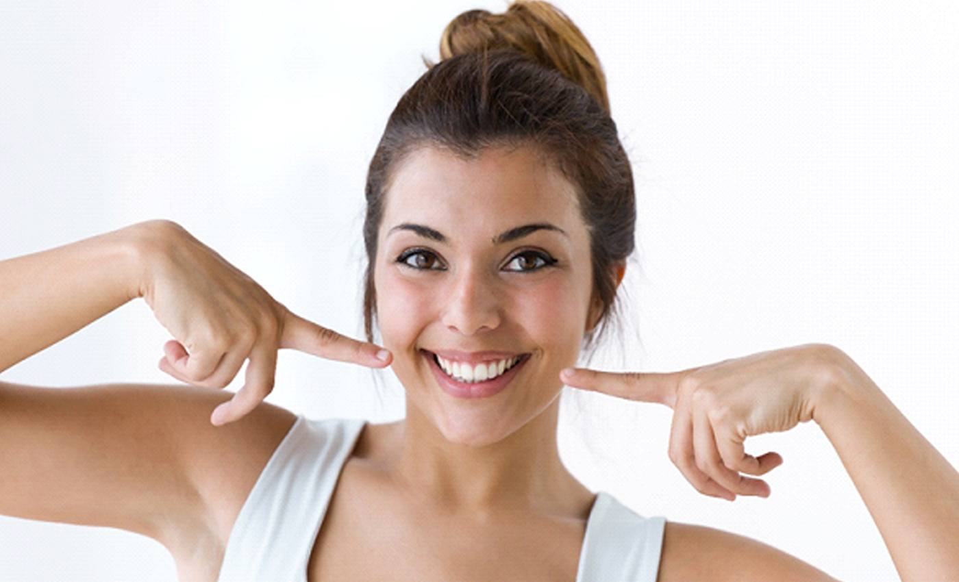 A young female wearing a white sleeveless blouse and pointing to her smile after receiving in-practice teeth whitening in Leesburg, VA