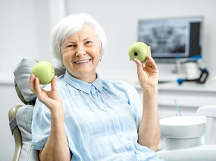 An older woman seated in the dentist’s chair holding two apples and smiling 