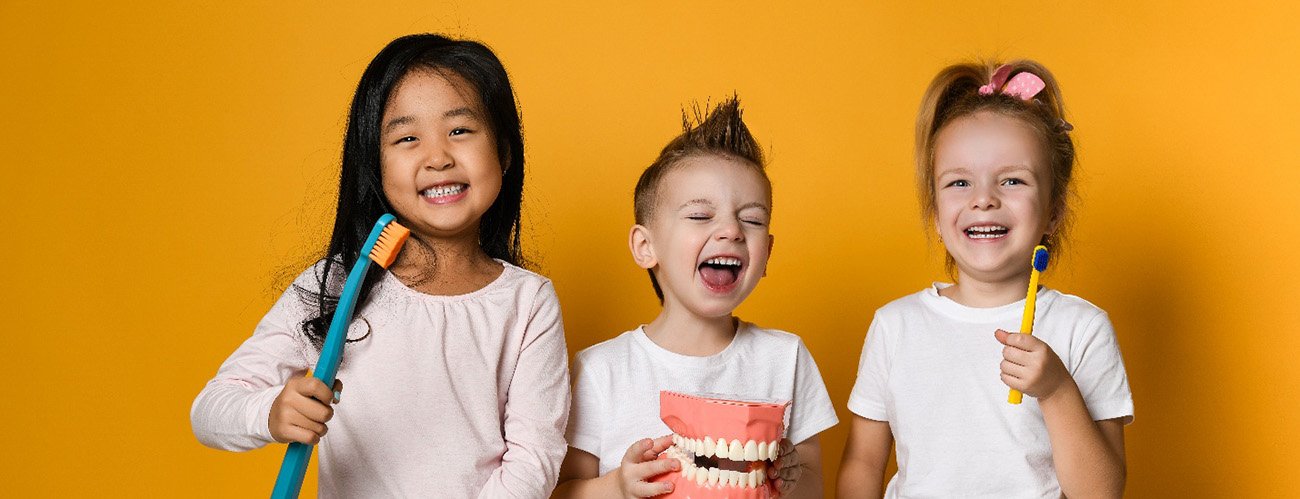 3 Kids holding oversized toothbrushes and model teeth smiling with goldenrod background