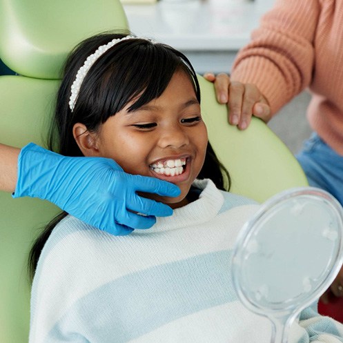 Dentist in blue gloves touching chin of girl in dental chair looking into mirror