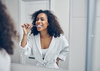 A curly-headed woman brushing her teeth in front of a bathroom mirror