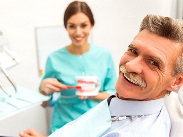 An older man smiling after receiving his dental implants and his dental hygienist showing him how to properly care for them