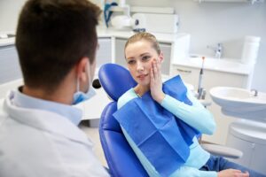 Patient in dentist’s chair holding face in pain, talking to dentist.