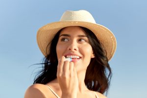 Woman with brown hair in a sun hat putting lip balm on in front of a light blue sky background
